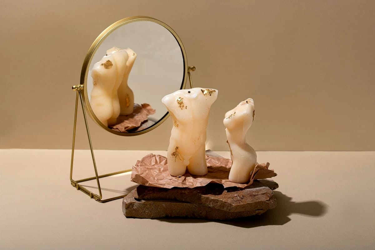 Two small sculptures of a male and female body, reflected in a nearby mirror that distorts and merges their forms, blurring the distinction between genders.