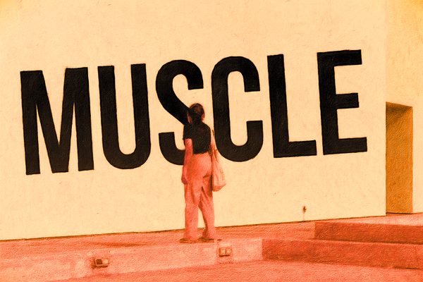 A woman in a museum gazes thoughtfully at the word “Muscle,” written in bold, prominent lettering across a gallery wall.