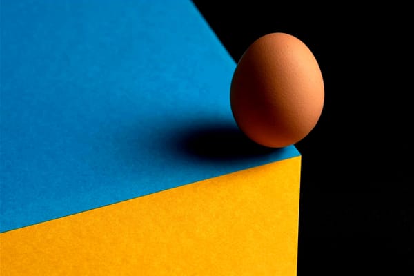 A single egg balanced precariously on the sharp edge of a vibrant, multicolored structure, teetering as if about to fall and crack.